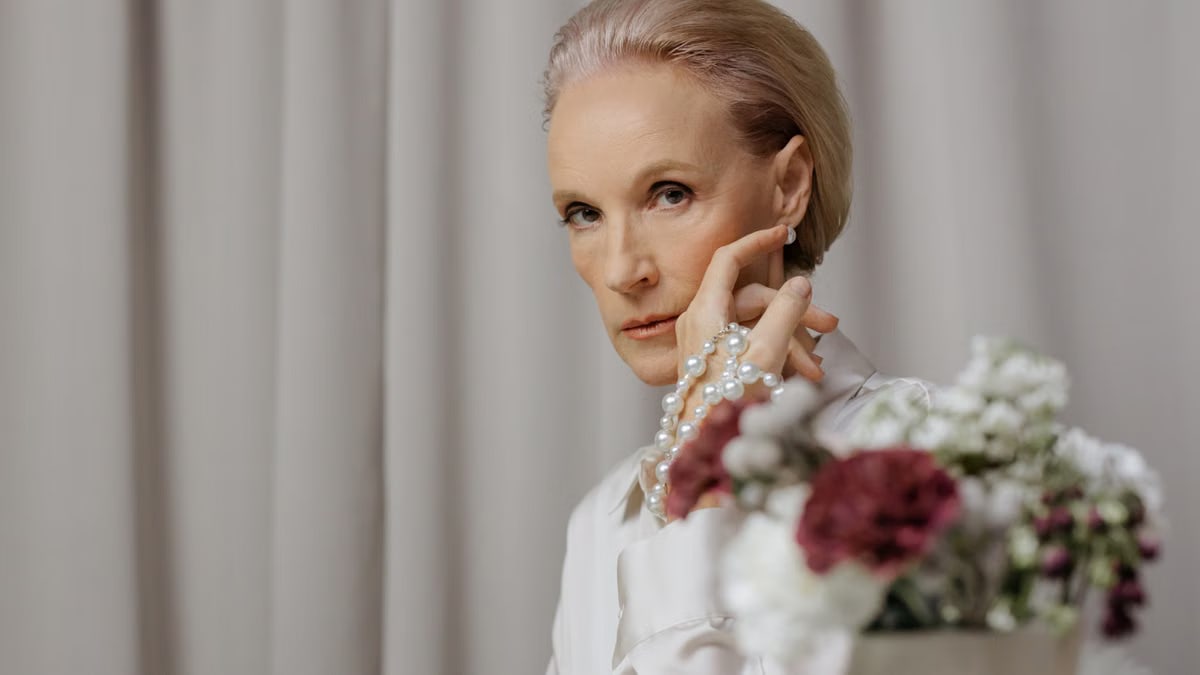 Elegant senior woman adorned with pearls and floral arrangement indoors.